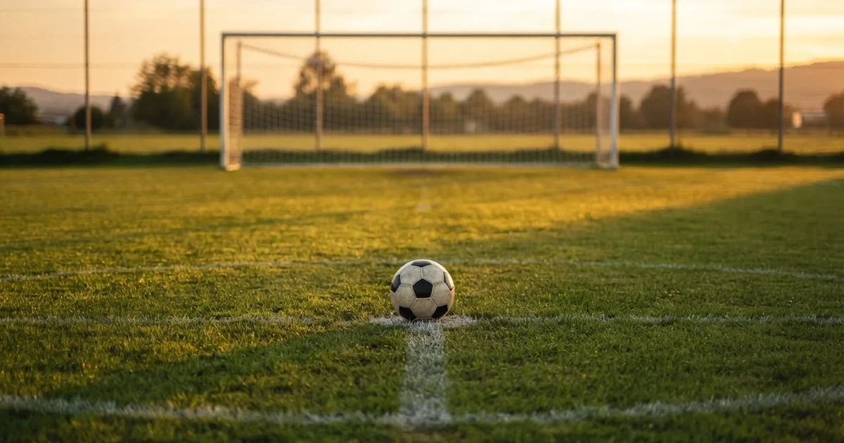 Ein Fußball liegt vor einem leeren Tor auf dem Rasen, das Netz hängt unbewegt, im Hintergrund ein verschwommener Zaun in warmem Abendlicht.