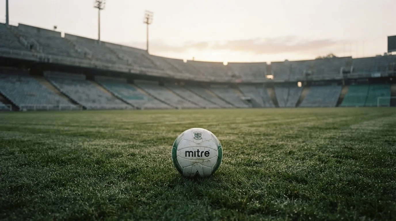 Ein Fußball liegt auf dem Rasen vor einer leeren Tribüne am frühen Morgen, Tau glänzt auf dem Gras.