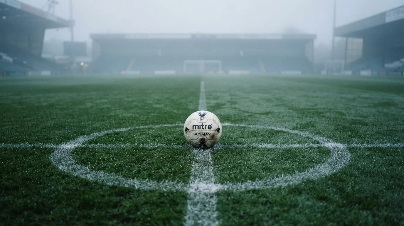 Ein Fußball liegt auf dem Anstoßpunkt eines leeren Rasenplatzes im Morgennebel.