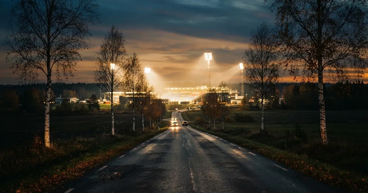 Ein Stadion unter Flutlicht aus großer Entfernung gesehen, davor eine dunkle Landstraße und einzelne Bäume in der Abenddämmerung.