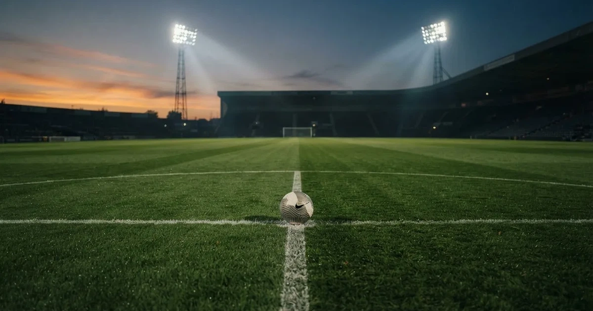 Ein Fußball liegt auf dem Anstoßpunkt eines Rasenplatzes in der Dämmerung, in der Ferne leuchten Flutlichter, die Ränge sind leer.